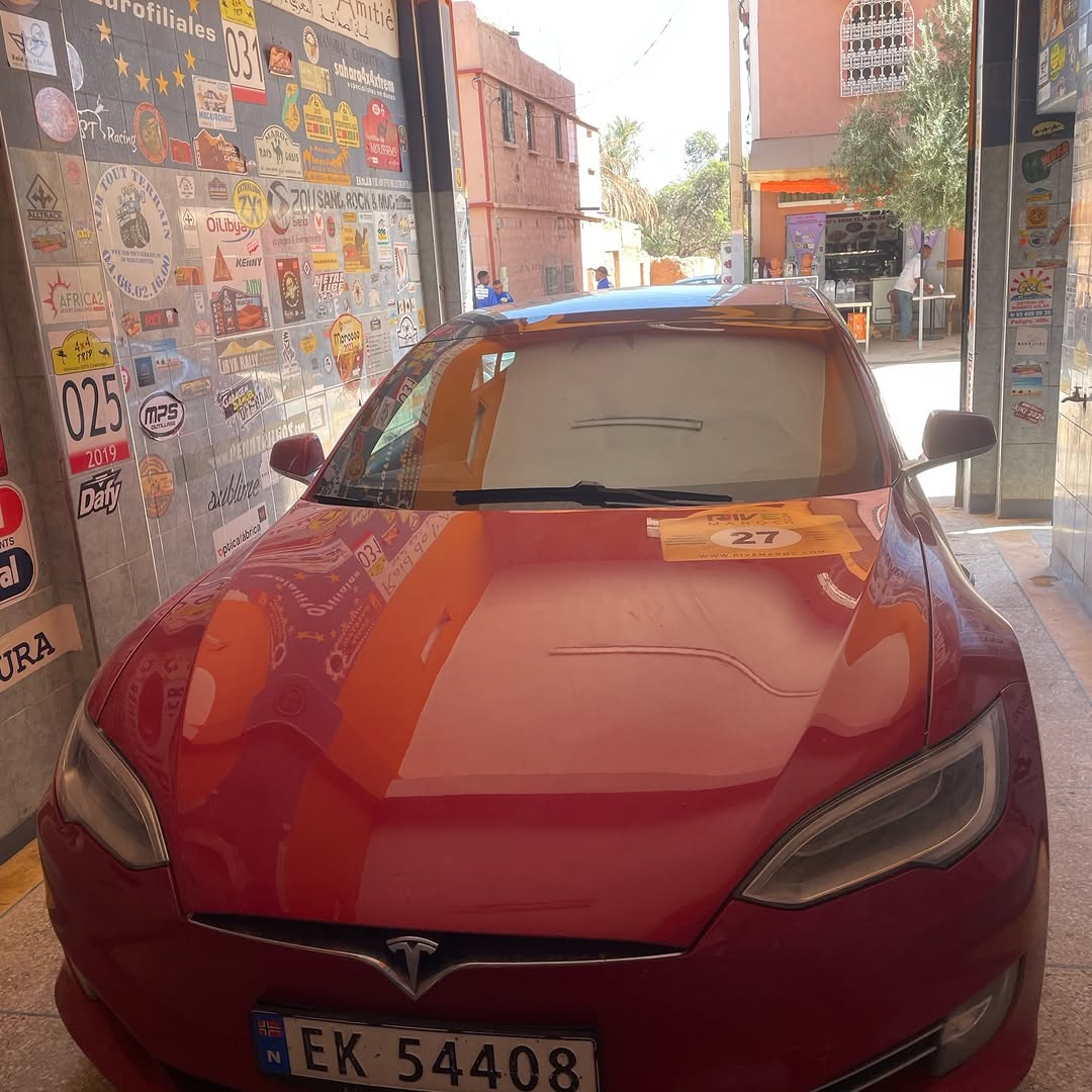 Red Tesla Model S with Rive Maroc rally plate 27 parked in the workshop entrance at Garage Iriki after the lift
