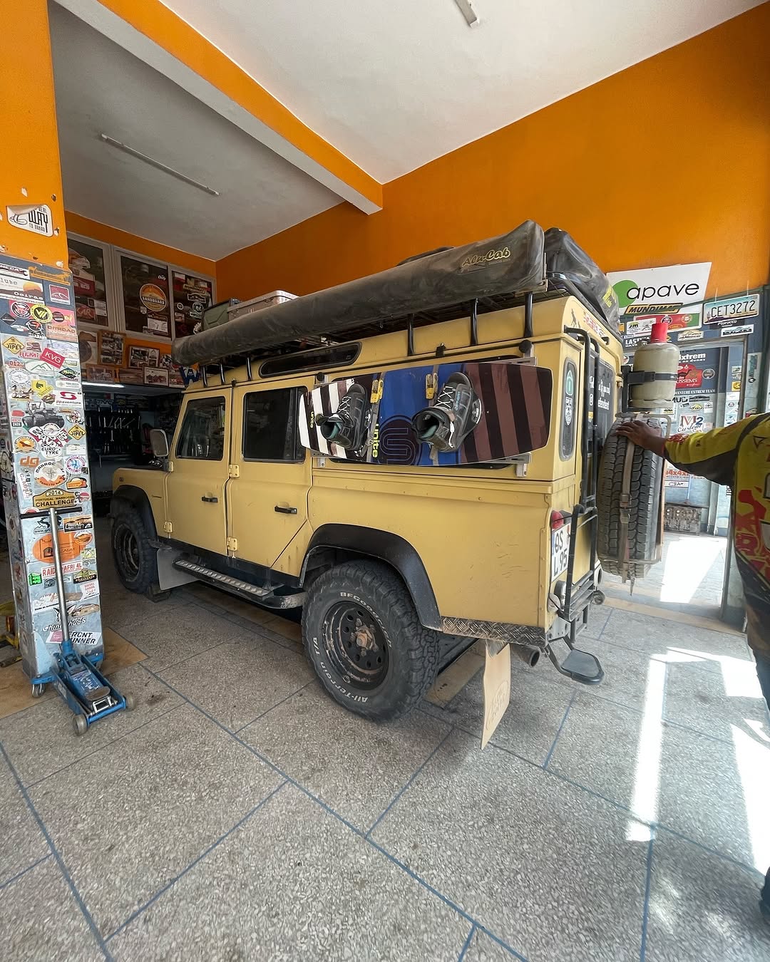 Rear view of the yellow Defender 110 with roof tent and snowboards mounted on the side windows inside the workshop